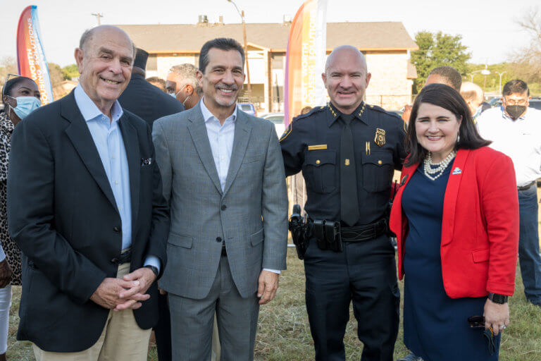 A group of four people, including a police officer and individuals in business attire, stand smiling outdoors with others in the background.