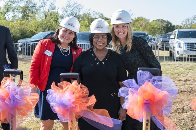 Three women in hard hats with orange and purple ribbons on shovels stand outdoors at a groundbreaking event.