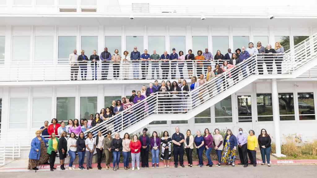 A large group of people stand in lines on a staircase and in front of a multi-story white building, posing for a group photo.