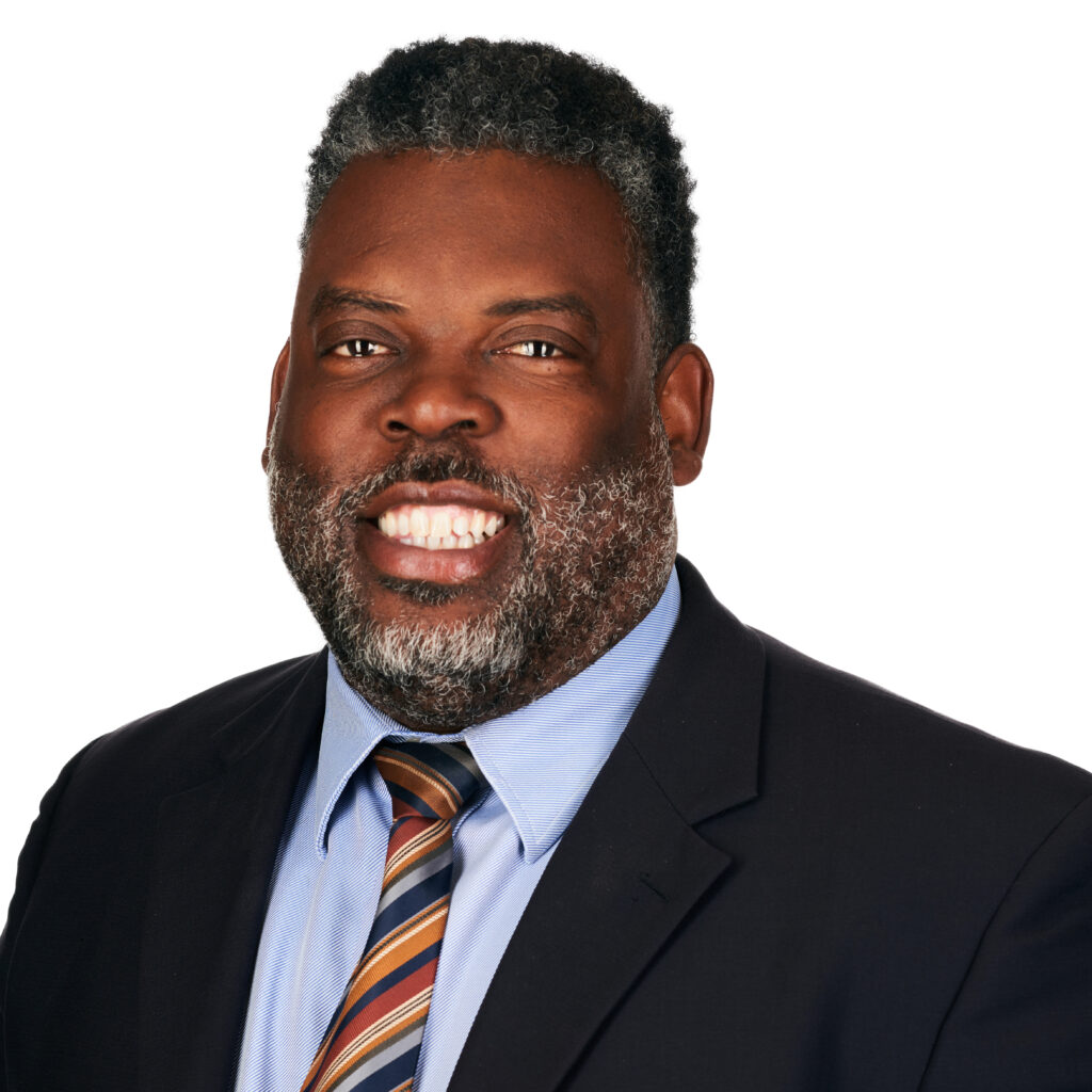 Smiling man with a beard wearing a plaid suit jacket and blue shirt against a gray background.