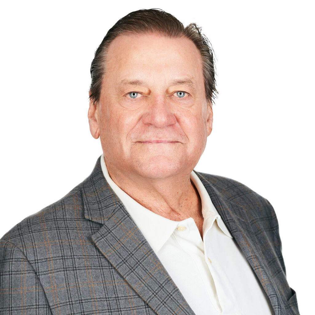 Portrait of a smiling man with short brown hair, wearing a checked blazer and yellow shirt, set against a gray background.