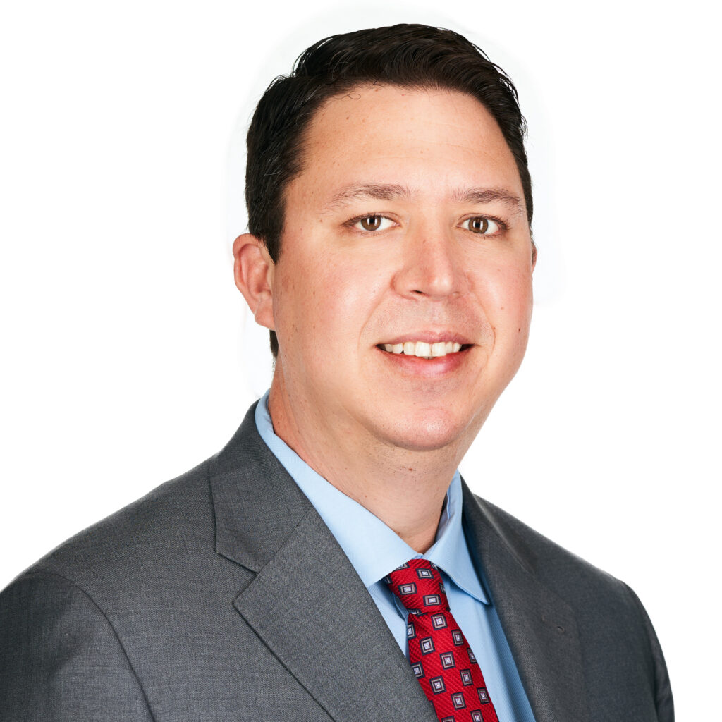 A person in a suit and tie smiles at the camera against a neutral, textured background.